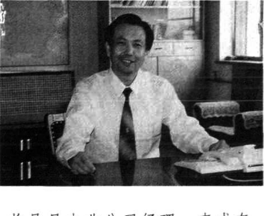 一张黑白照片，展示一名男性坐在办公桌前微笑，桌上摆放着办公用品和设备。