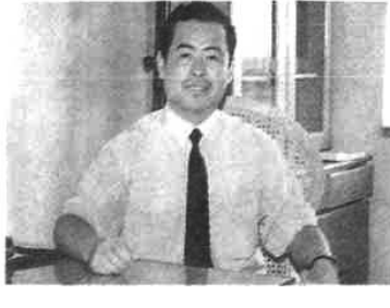 黑白照片，显示一位身穿白衬衫系领带的男子坐在室内办公桌前。