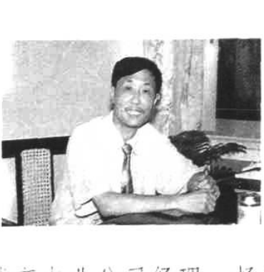 一张黑白照片，显示一位身穿短袖衬衣、系领带的男子坐在桌前微笑。