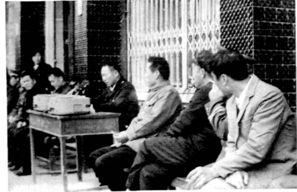 一群人在室内，其中几人坐在桌子后面，一人在讲话。