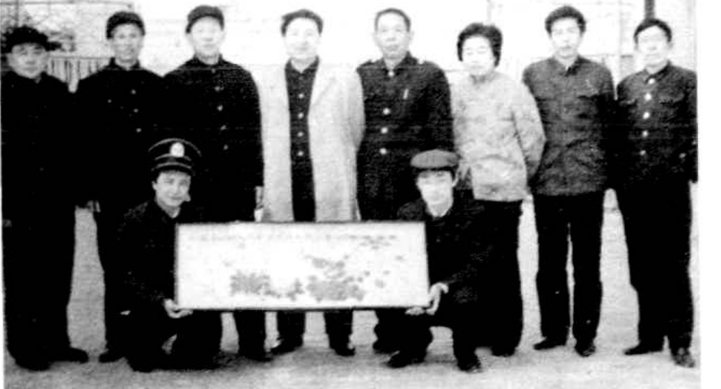 一群身着深色服装的 men 和 women 站成一排，其中两人向前一步，手中拿着一幅画