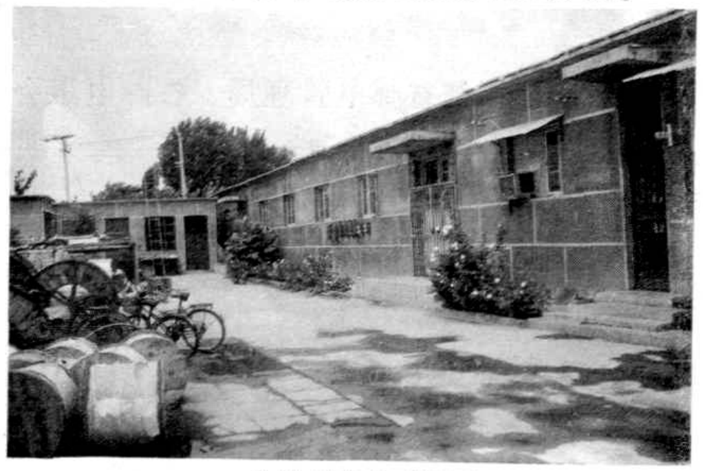 一排平房式建筑，前方有水泥地面，地面上有堆放的圆柱形物体和自行车。