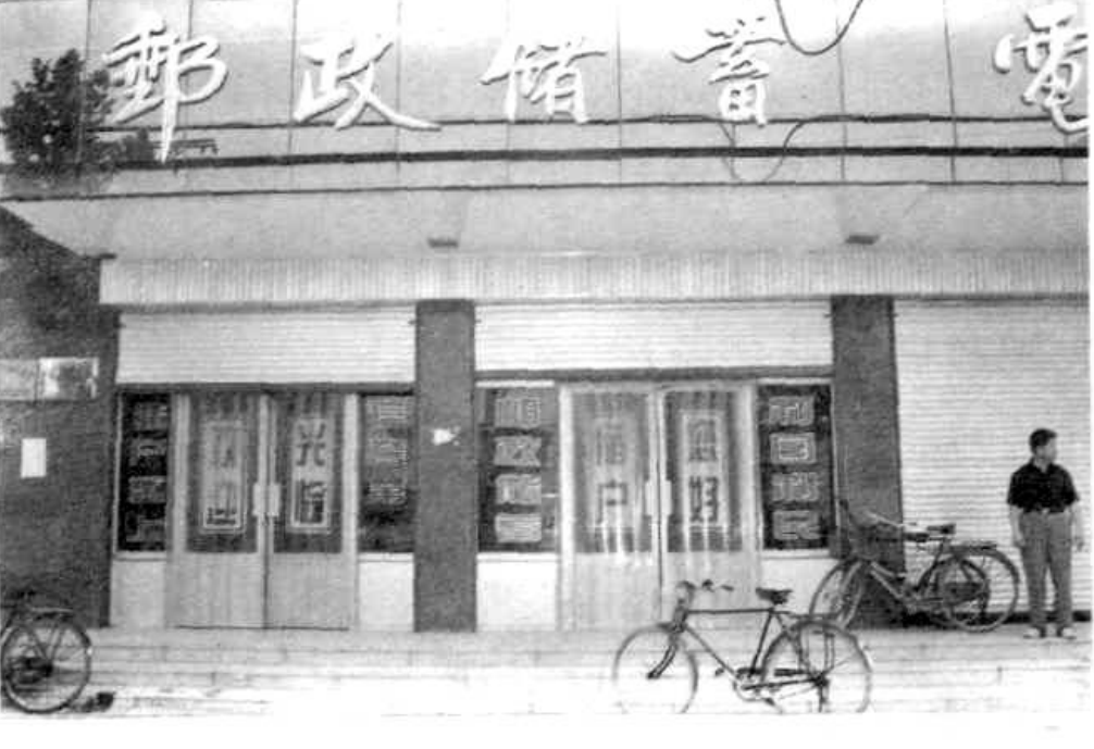 一间带有“邮政储蓄”字样的建筑的外景，前面停放着两辆自行车，左边还有一个男子。
