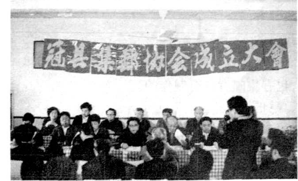 一张合影照片，一群人坐在桌子后面，背景有写着“冠县集邮协会成立大会”的横幅。