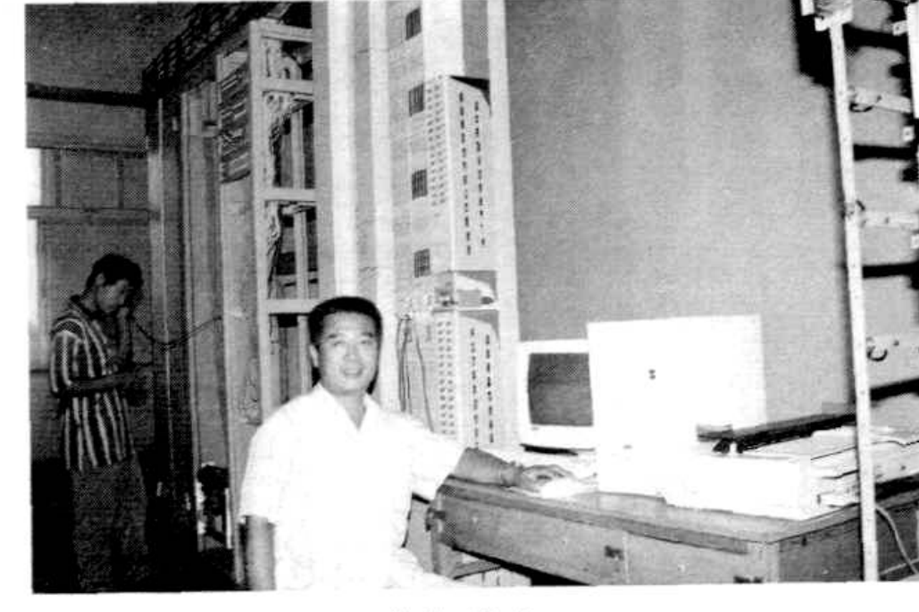 两名男子站在一间满是电信设备的机房内，其中一人正在操作一台显示器。