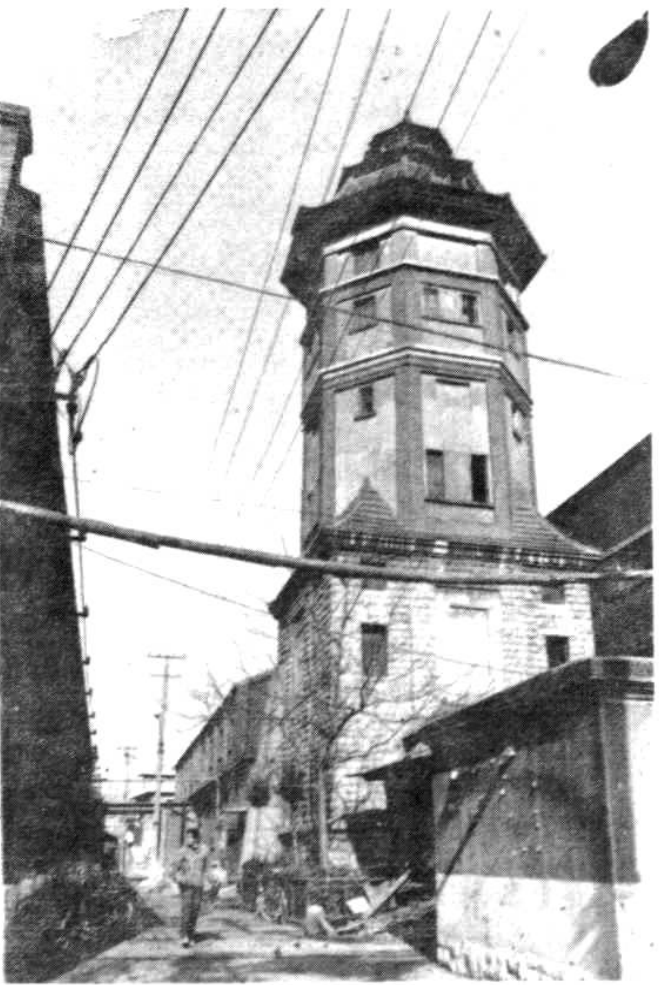 一张黑白照片，展示一座建于1910年的津浦铁路济南机务段百吨水塔，水塔为多边形砖石结构，带有窗户，周边有低矮建筑和街道设施。