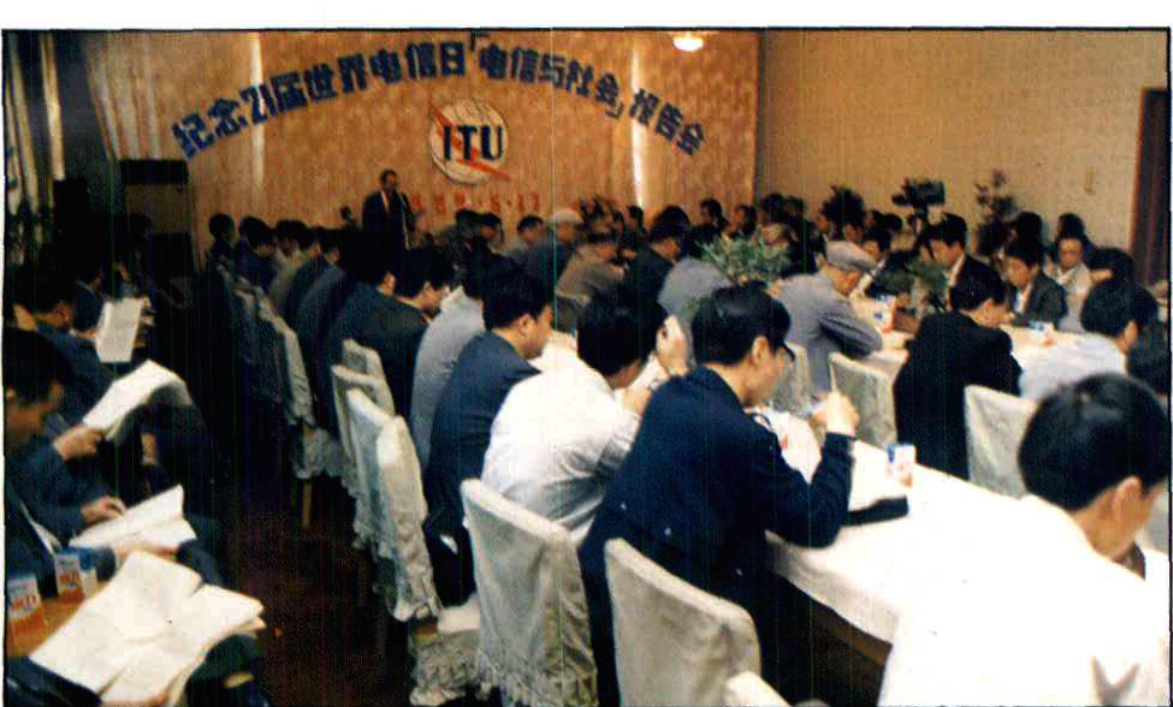 室内会议场景，多排参会人员坐着听会，前方有演讲台和背景横幅