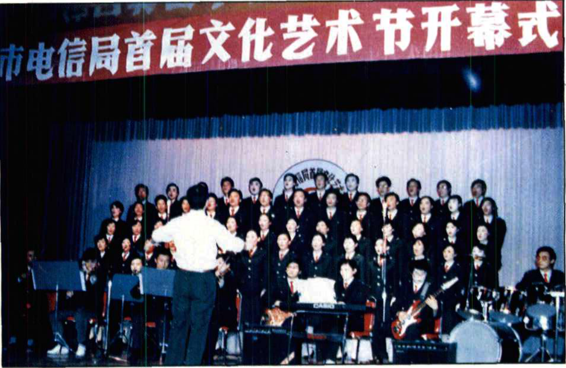 舞台上合唱团正在表演，前方有乐队和背对镜头的指挥，背景挂有横幅