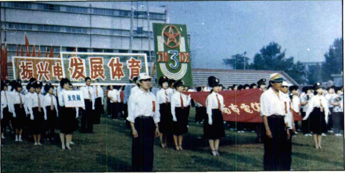 户外草坪上身着制服的方队和人群，背景有标语牌写着振兴发展体育