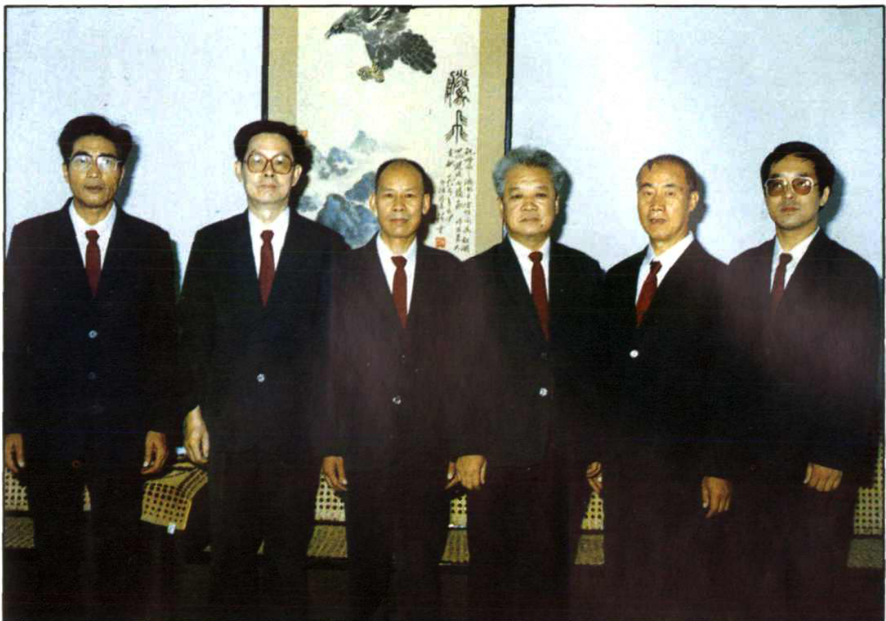 六名身着西装的男性站在室内合影，背景是一幅字画。