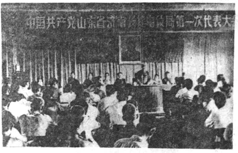 黑白历史照片，展示一处室内会议现场，背景挂有横幅，聚集了许多参会人员。