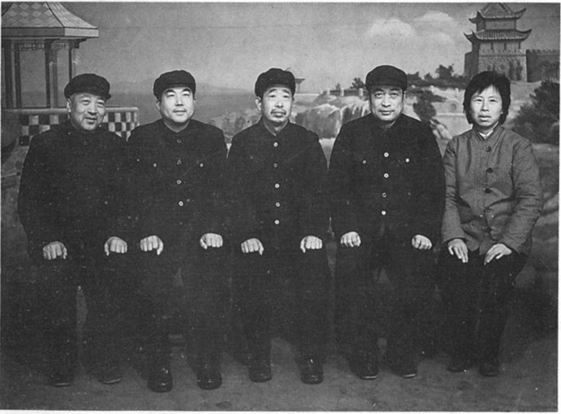 黑白合影照片，五位穿着中山装或夹克的工作人员在室内布景前合影，背景带有亭台楼阁绘景。