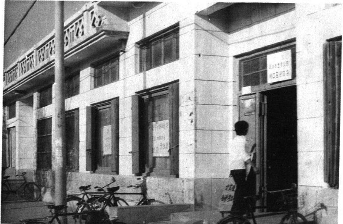 黑白照片，展示一处零售门市部的沿街建筑外景，门前停放着自行车，有人正在出入。