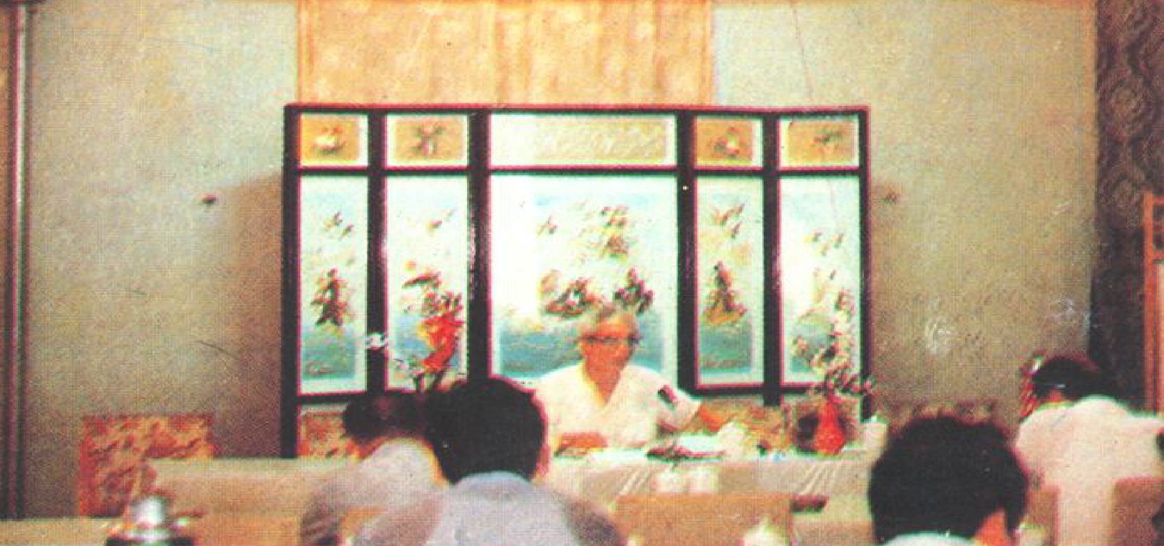 在会议室里，人们围坐在桌子旁，桌上摆放着文件和饮品。背景墙上挂有写有中文的横幅，中间有屏风隔断。