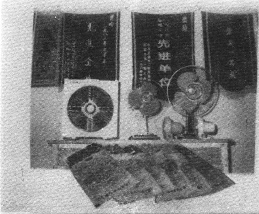 黑白照片，展示家用电器厂生产的电风扇、电热褥和背景中的锦旗。