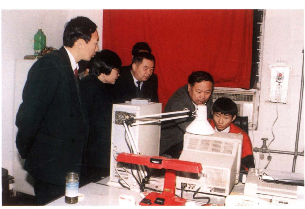 几位身穿西装的工作人员在一台电脑和桌子前观察讨论，身后是红色背景墙