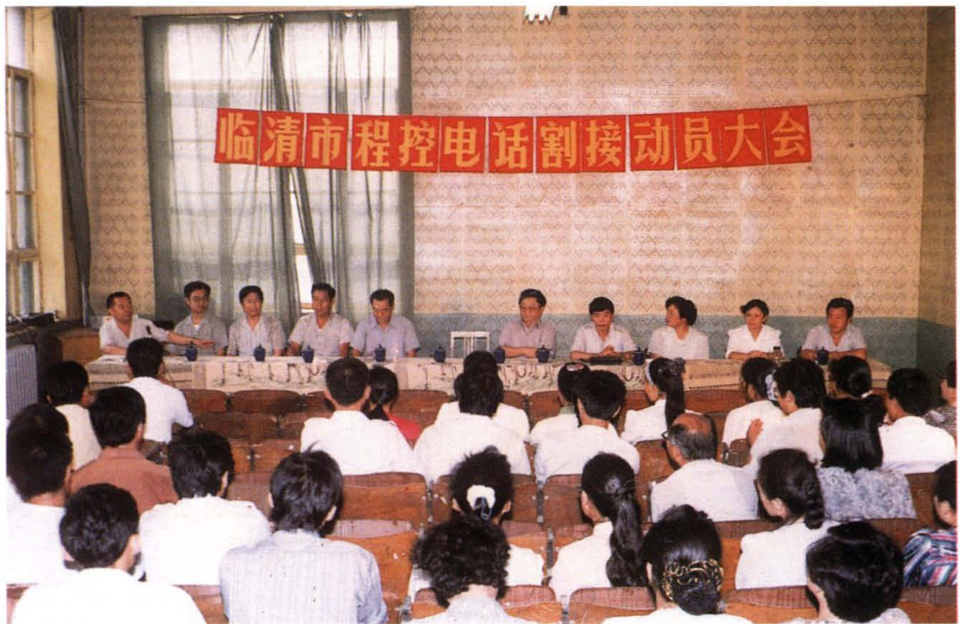 一大会议室场景，前方主席台坐有多人，墙上有“临清市程控电话割接动员大会”横幅，台下坐满参会人员的背影