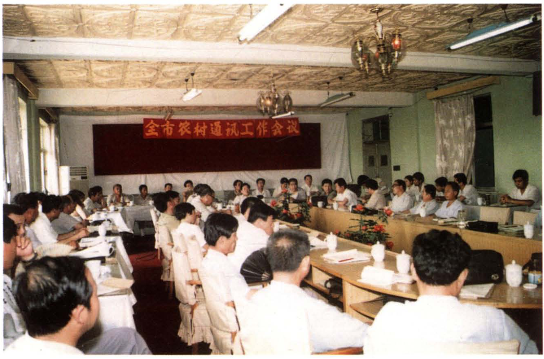 会议室内部场景，长桌围坐着众多与会人员，墙上挂有“全市农村通讯工作会议”红色横幅
