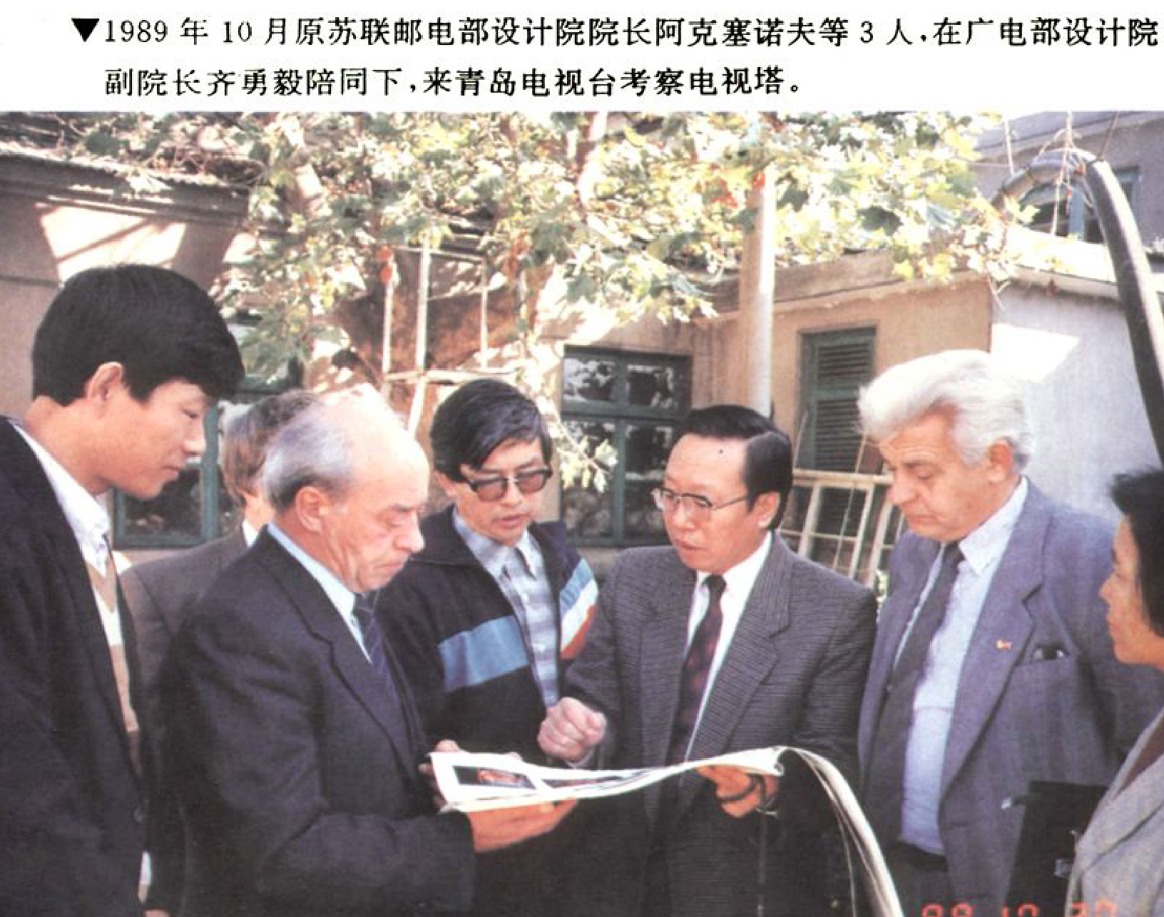 一群人在室外查看文件，其中一位年长男士拿着文件，周围的人围观。