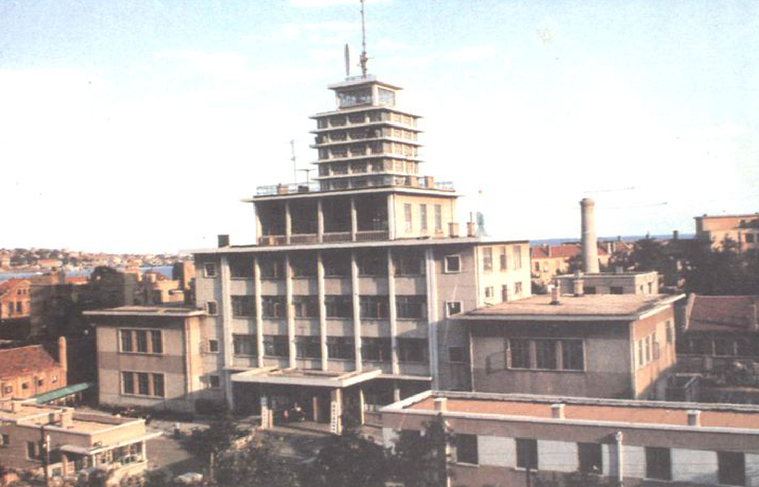1980年建成的一座多层建筑，顶层有塔楼，远处有城市和港口景观。