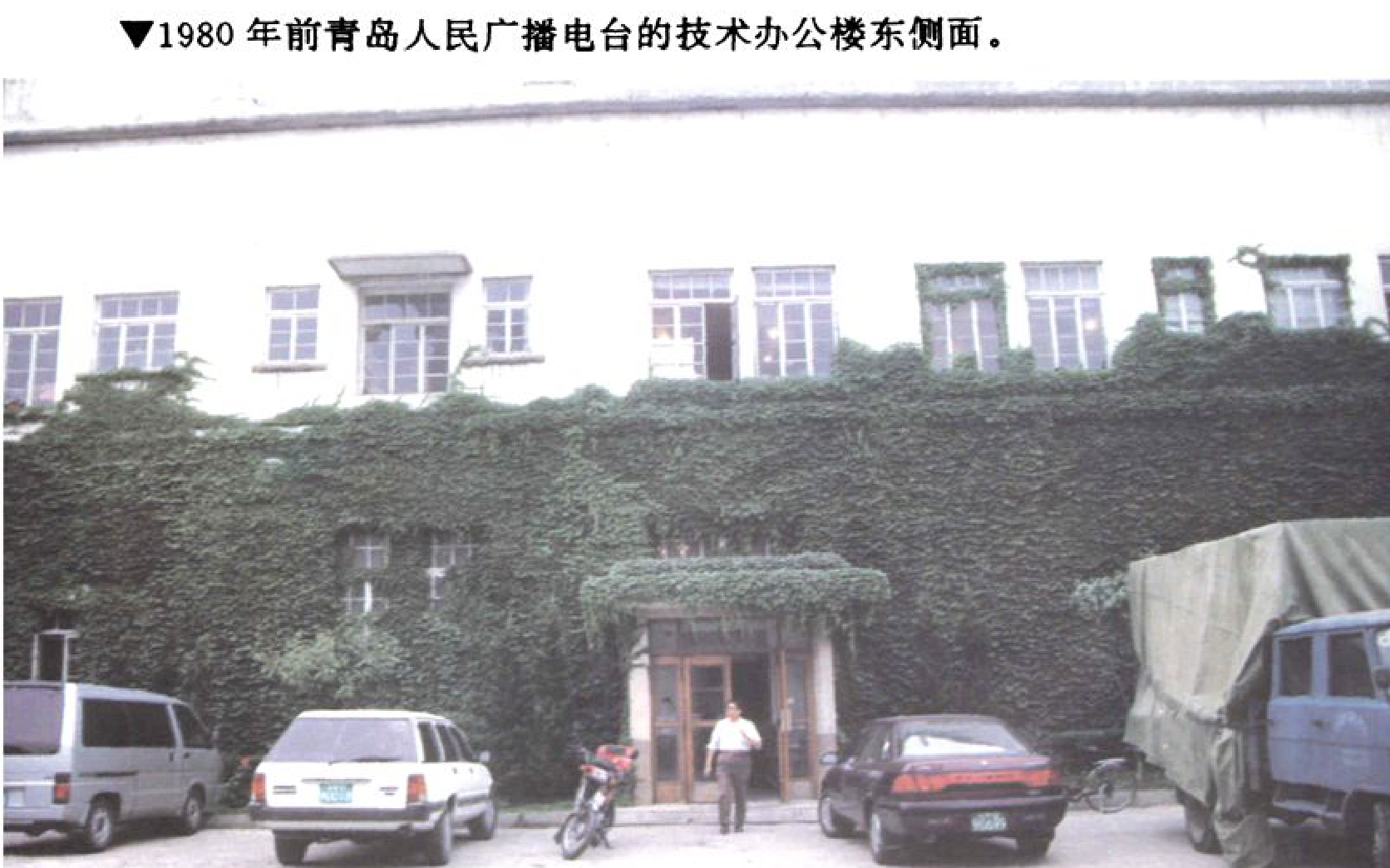 一栋被爬山虎覆盖的白色建筑，入口处停有汽车、摩托车，旁边有行人。
