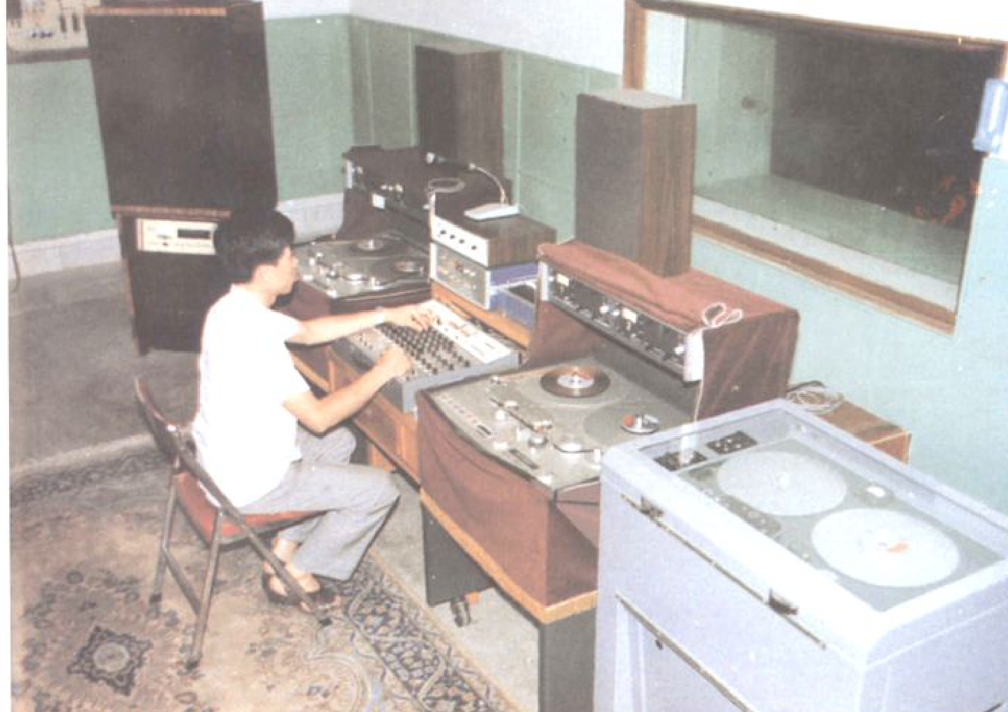 一个人坐在控制台前，操作着录音设备，房间内有多个扬声器和录音机。