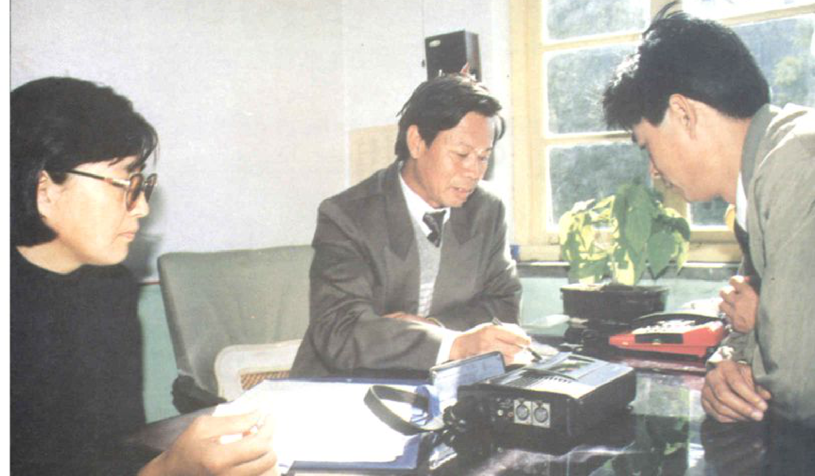 三个人围坐在一张桌子旁，桌上放着录音设备，两人正在交谈。