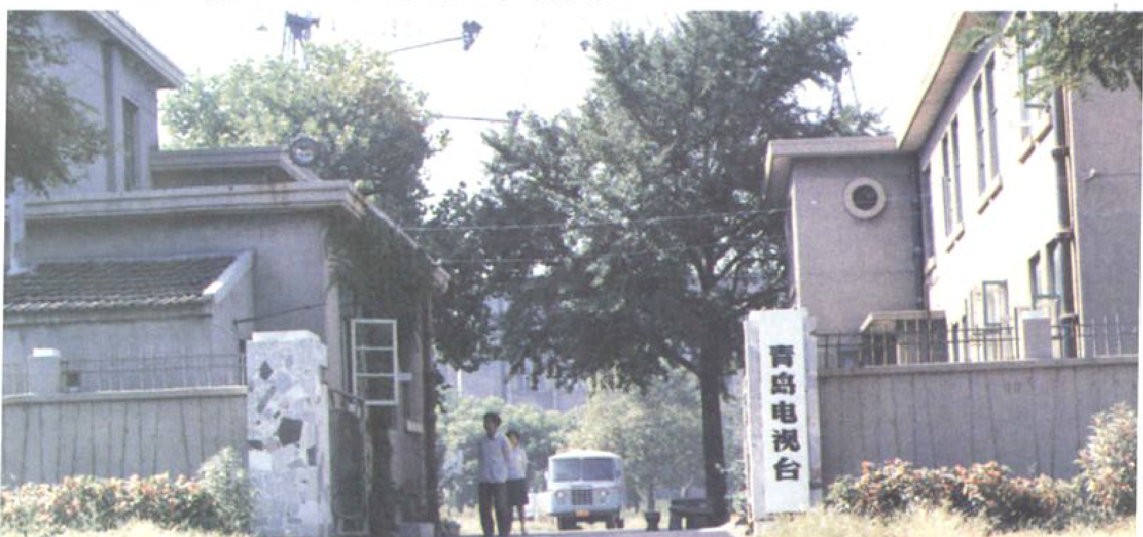 一条道路通向一栋建筑，路边有门柱和牌匾，有两人和一辆面包车。