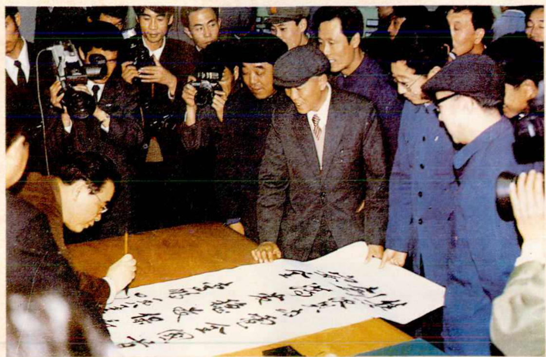 一行人围在一张桌子旁，其中一人正在一张展开的长纸上写毛笔字，其他人围观并拍照。