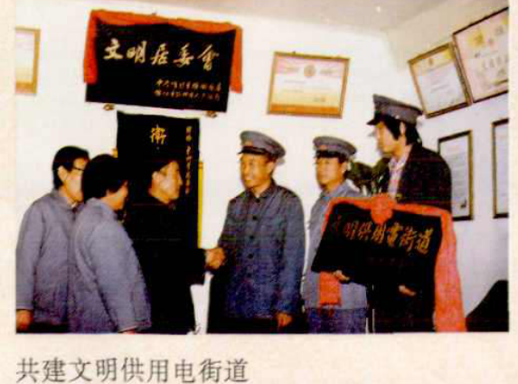 几个人站着，其中一人在向另一人展示一幅写有“文明供用电街道”的旗帜。