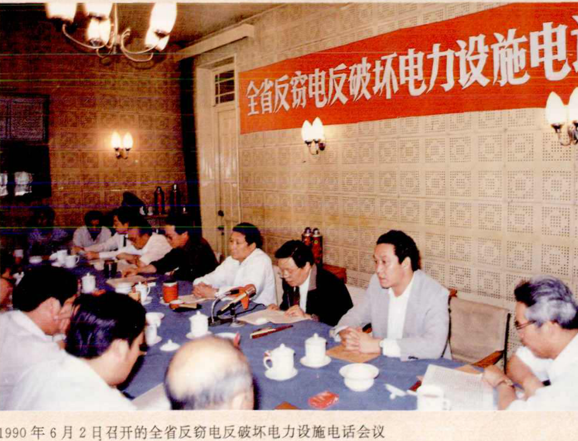 一群人围坐在一张长桌旁开会，桌上有茶杯和文件，墙上挂着红色横幅。