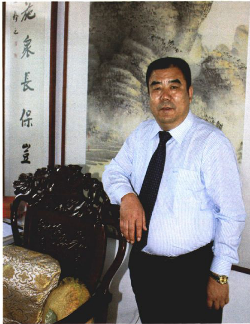 一名中年男子站在一张椅子旁边，身穿浅蓝色条纹衬衫和深色领带，背景是一幅画作和一副书法对联。