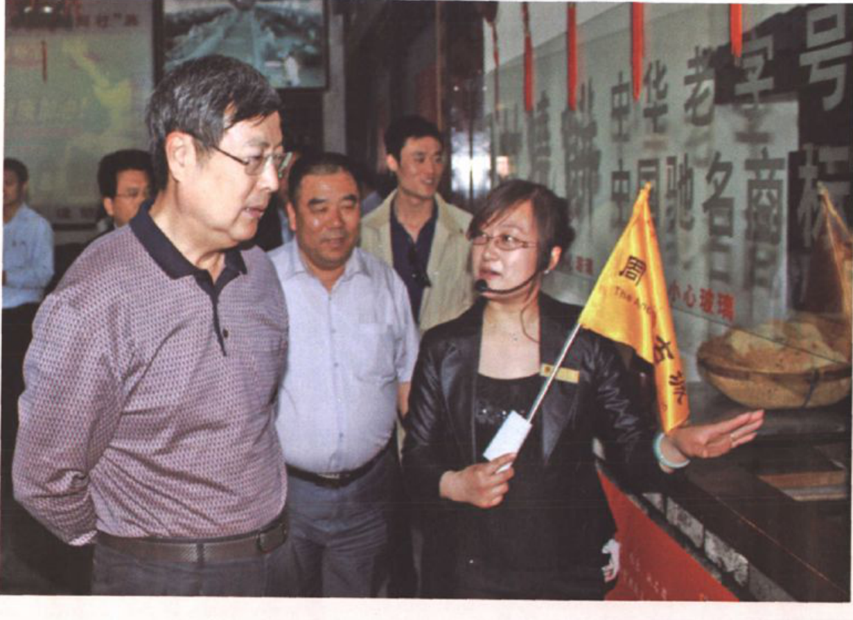 多人参观室内场景，左侧戴眼镜男子注视前方，右侧女士手持黄色导游旗讲解