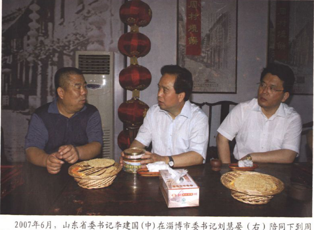 三位男性坐在桌前交谈，桌上有烧饼、茶具和纸巾盒，背景有串联的红灯笼和挂画