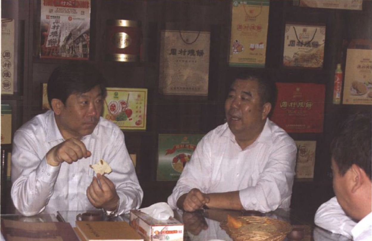 两名男子坐在室内桌前交谈，墙上摆放着各种包装和礼盒