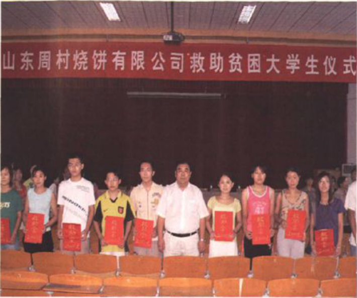 一张彩色合影照片，横幅写有“山东周村烧饼有限公司救助贫困大学生仪式”，多名学生和领导站在舞台上展示证书