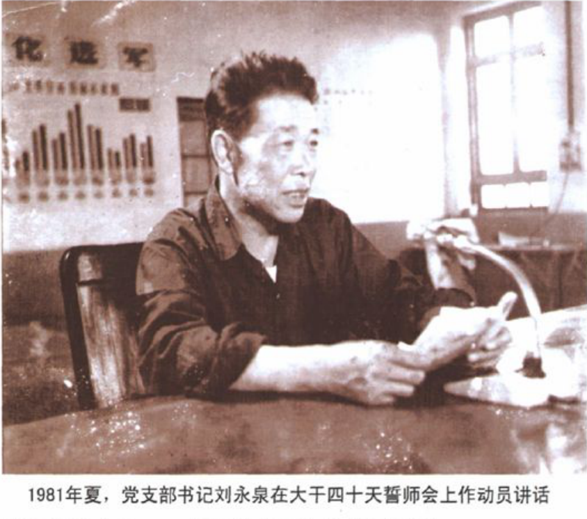 黑白照片，一名男子在室内手拿纸张讲话，背景是带有宣传标语和人群的场景