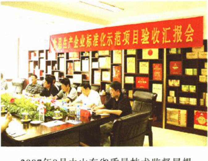 室内会议合影，上方横幅字样为“××品生产企业标准化示范项目验收汇报会”